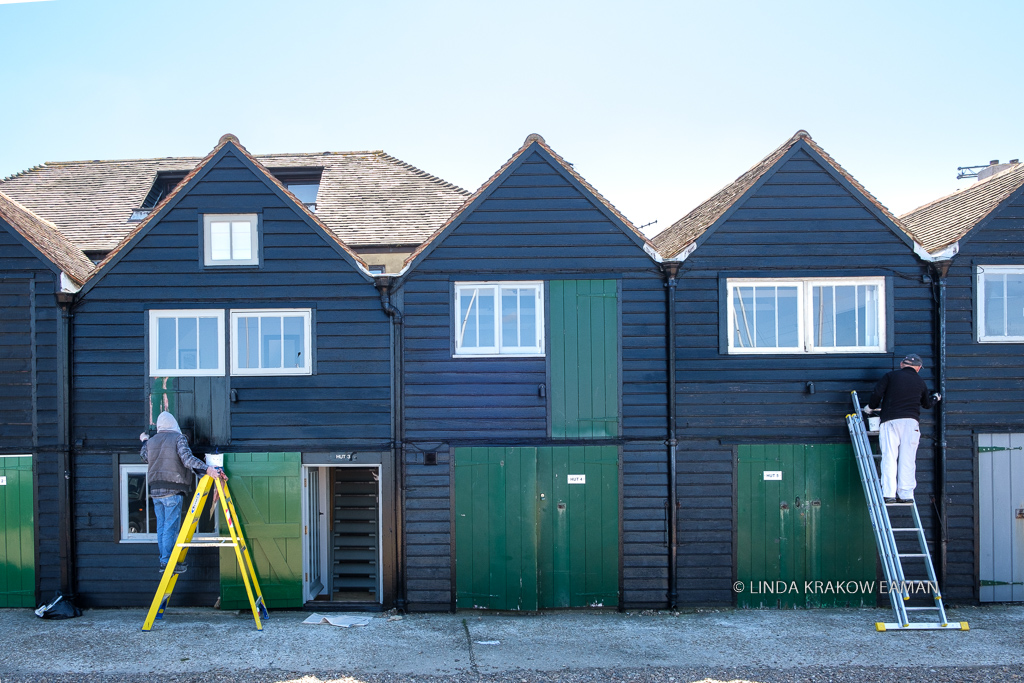 Row of three narrow, dark blue or black buildings with dark green doors. On each end is a person on a ladder, painting the building. 