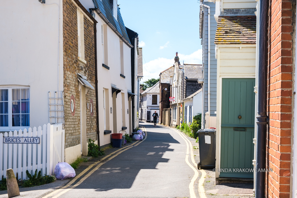 Narrow street with modest one and two storey houses on each side, with doors that  open directly to the street. A man walks down the street toward us, in the distance. 