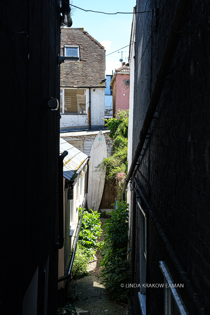 A glimpse of the narrow space between two buildings, with a white surf board leaning up against a wall at the end. 
