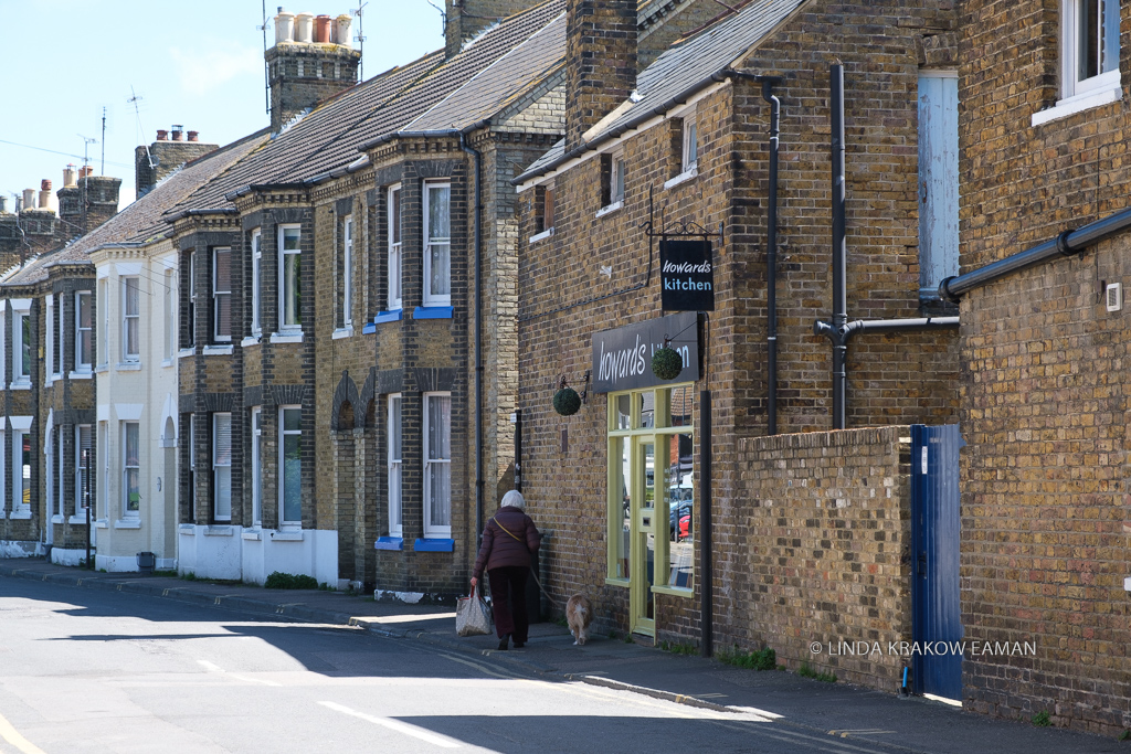 A row of brown brick buildings, a restaurant in the building closest. A woman walks her dog, carrying a shopping bag, on the sidewalk. 