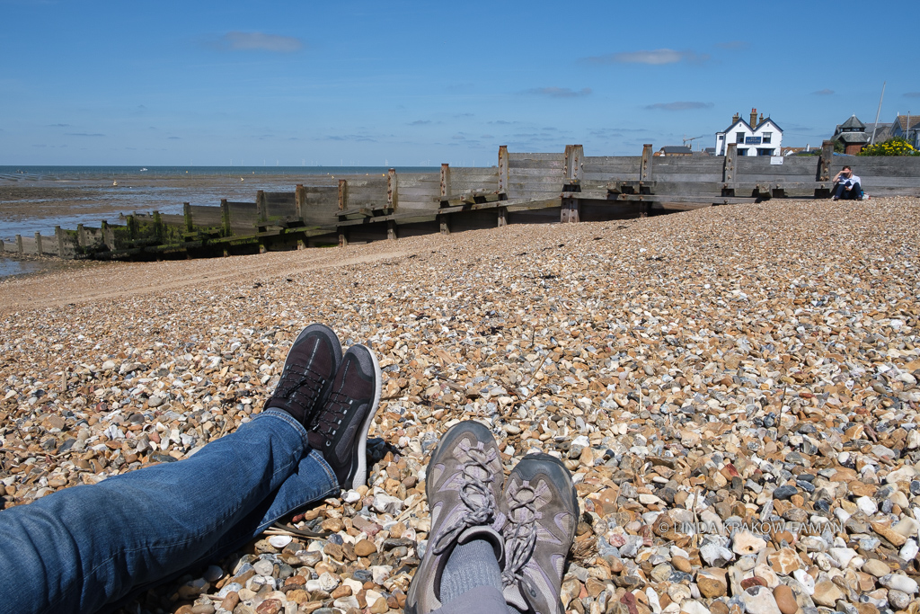 The photographer's feet, and her companion's, show they are sitting on a rocky beach. A groyne is in front of them, and beyond that a white house, the ocean, and a blue sky. 