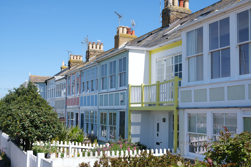 A row of white two-storey houses with lots of windows and pastel colored accents. White picket fences enclose small gardens. 
