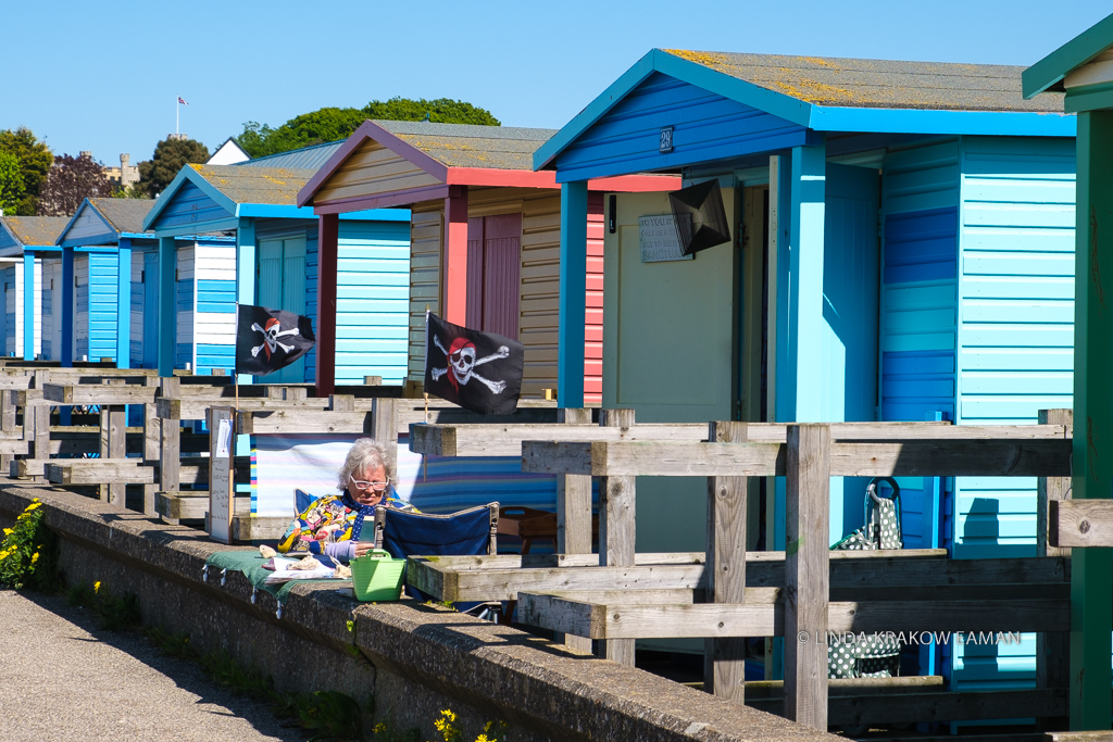 A woman sits reading in the tiny front yard area of her beach hut. The three of four nearby huts are all closed up for the winter. 