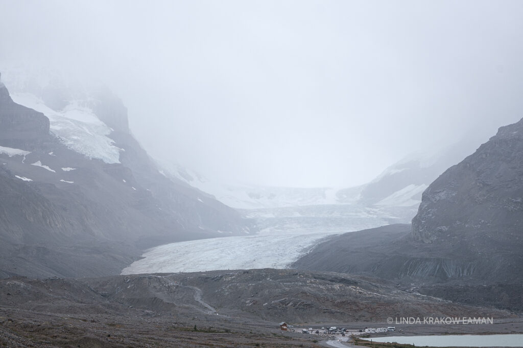 Ice flows between rocky hillsides. The sky is white with clouds and the buildings and cars in the parking lot below are tiny in comparison.