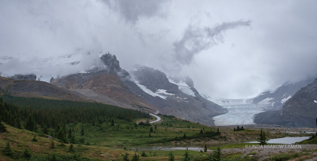 The glacier is now in the far left of the image; a green landscape is in the foreground with the mountains and glacier in the background. 