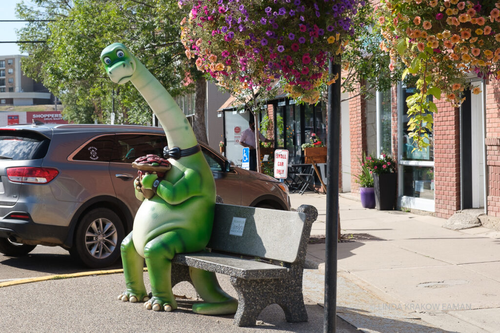 A cute green dinosaur statue sits on a city bench looking curiously at the viewer. Shops are visible in the background, and there are flowers in the foreground hanging from a post. 