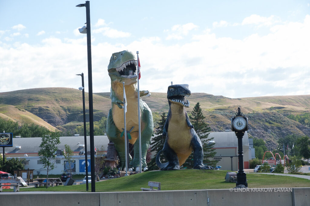 Two huge dinosaur statues sit on a little green hill and tower over a low building behind them.