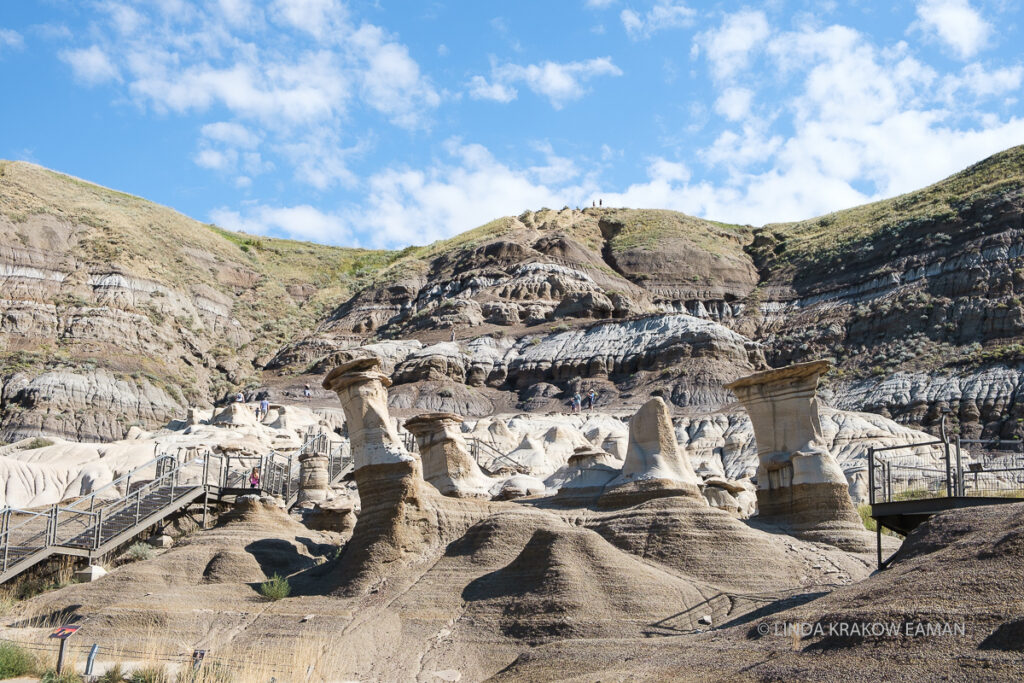 A blue sky over a butte, with five or six tall narrow columns of rock, called hoodoos, in the foreground. They are beige on top, brown on the bottom. A metal walkway with stairs winds around them. 