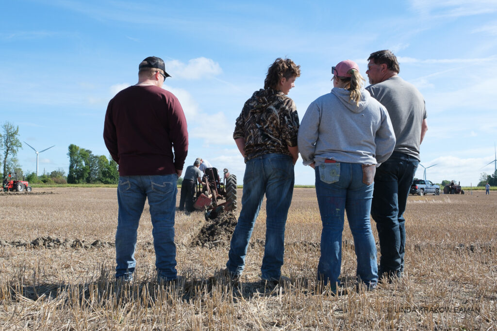 Four adults standing in a field of stubble, watching as a tractor starts plowing the field in front of them. 