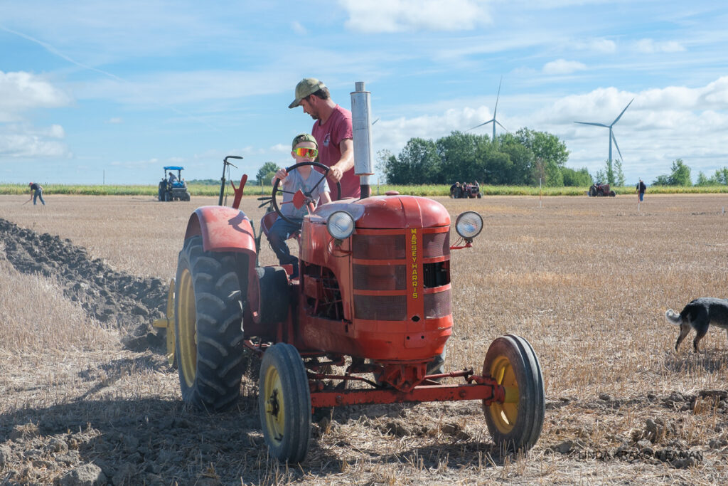 A six year old wearing reflective sunglasses sits at the wheel of a red tractor, his father standing beside him with one hand on the wheel and looking back at the furrow they have just plowed. 