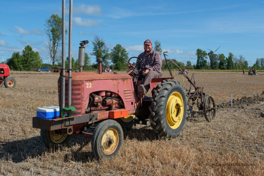 A smiling man in flannel shirt and cap is driving an antique red tractor pulling an antique plow. 