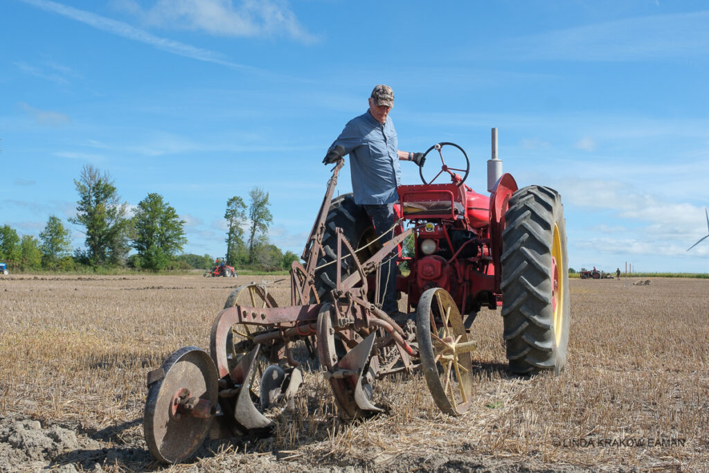 a man stands on the platform of a red tractor, facing behind it, with one hand on the lever that controls the furrows in his antique plow. 