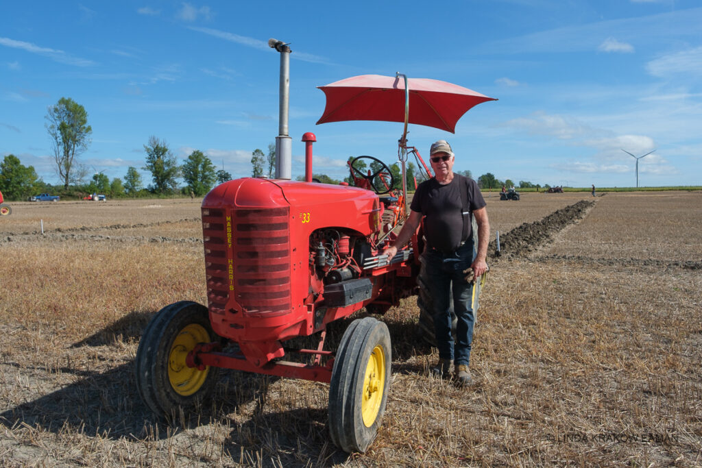 A man with stands in a field of crop stubble,  in front of an antique red tractor with a red umbrella over its seat. 