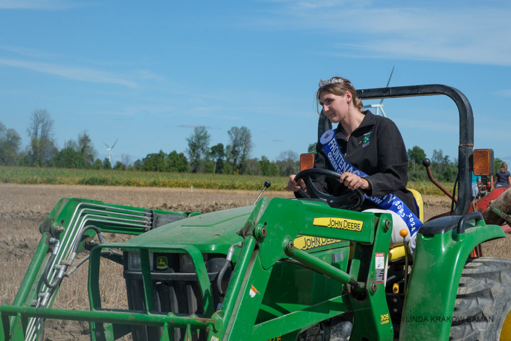 A woman wearing a tiara and blue sash sits on a John Deere tractor