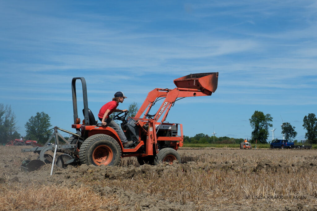 A boy in a red shirt leans forward in the seat of an orange tractor with front shovel lifted in the air, pulling a plow. 