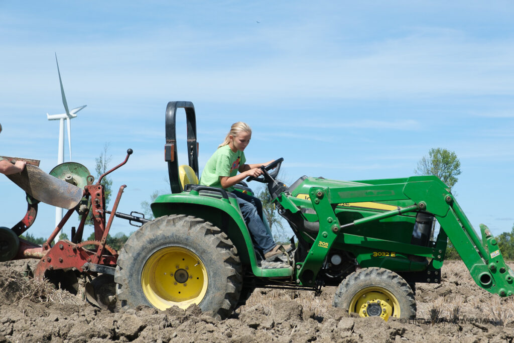 A girl in green tshirt operates a green tractor, looking down at the plowed ground beside her. 