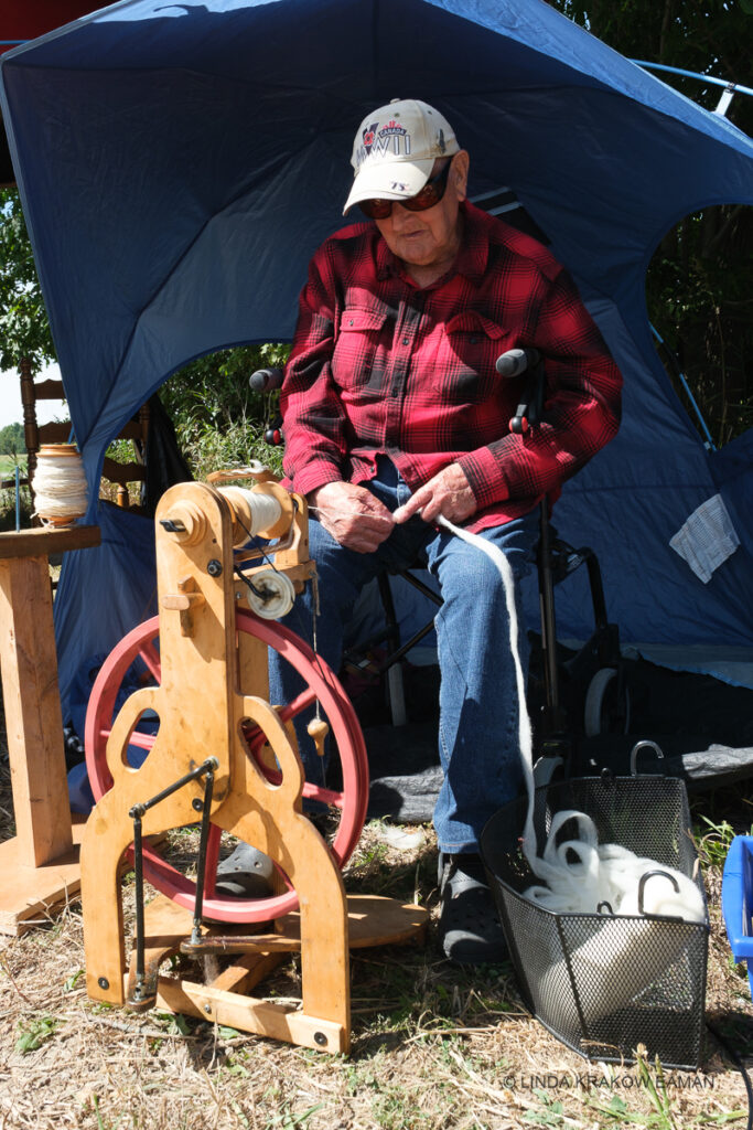 An elderly man wearing a WWII baseball cap sits under a blue sun shelter operating a small spinning wheel. 