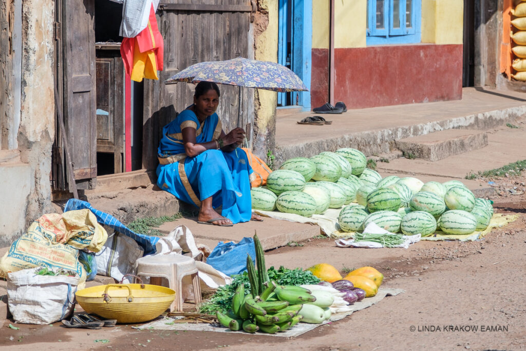 A woman in a blue and gold saree, with flip flops on her feet, holds an umbrella to shade her body. She is selling watermelons and some vegetables that are all laid out on cloths on the ground. She's sitting on a stoop, up against an old building. 