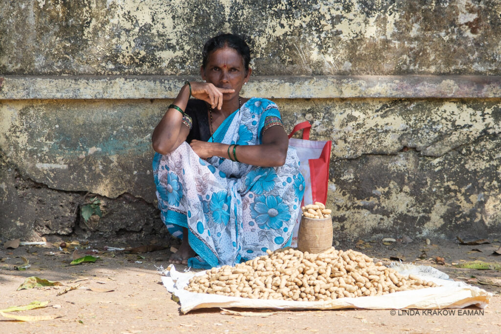 A barefoot woman in a blue and white flowered saree is squatting in a tiny patch of shade against an old stone or cement wall. She has a pile of peanuts on a cloth on the ground, with some piled in a cup. She is looking directly at the camera with a very serious expression. 