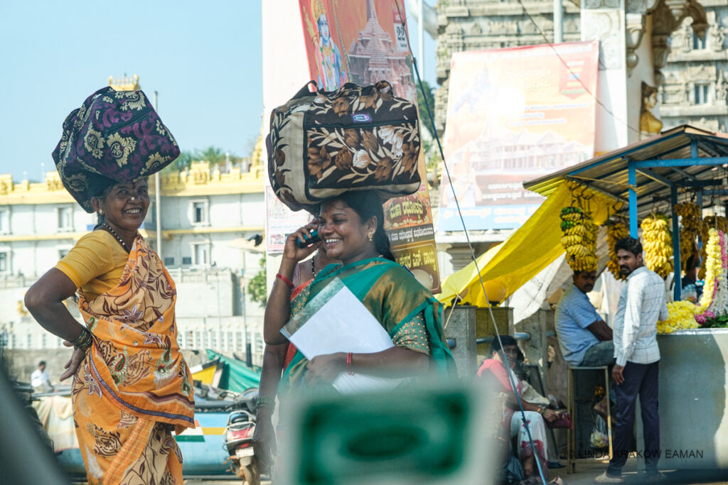 A messy shot through a car window: One woman in a yellow saree is carrying a soft sided printed bag on her head, smiling at the camera. Another woman in a green sari, with a similar bag on her head, is walking and smiling with a cell phone to her ear. Two men at a banana and flower stand are turned to look toward the camera. 