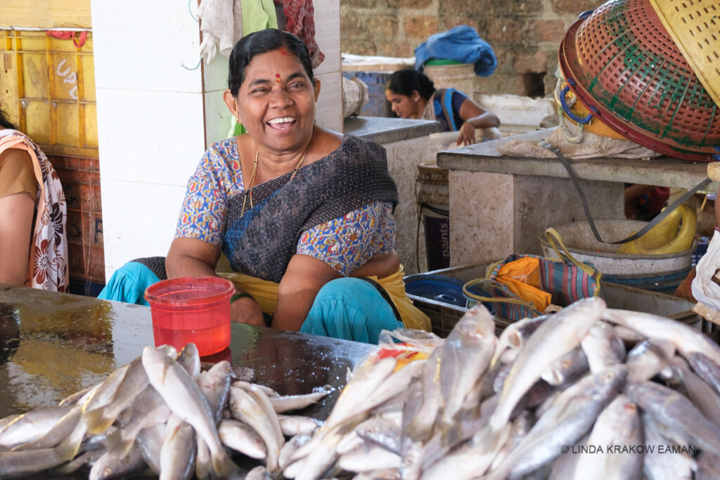 A seated woman laughs and looks at the camera. SHe's wearing a multi-coloured saree and in front of here is a pile of fish on a wet counter. Around her are baskets and other market items. 