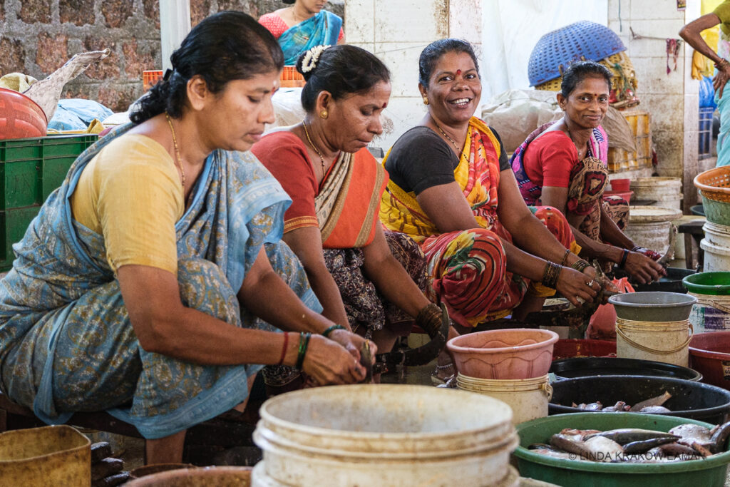 Four women in sarees sit low on upturned pails, their hands low in front of them as they clean fish. Two of them are looking at the camera and smiling