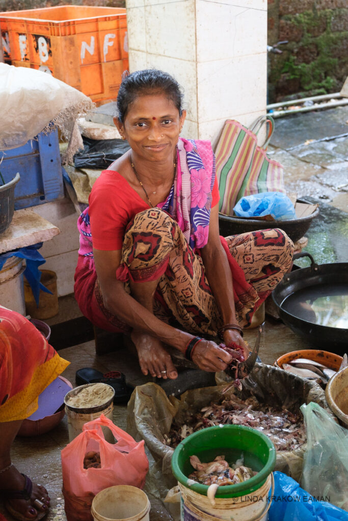 A woman in a saree, sitting low to the ground, looks up at the camera as she cleans fish.  There is a silver toe ring on the second toe of her bare foot. 