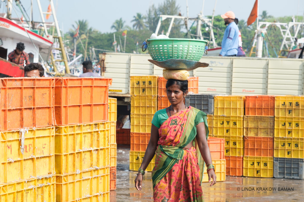 A woman in a colourful saree is carrying a green plastic basket full of crushed ice. She's walking past stacks of yellow and orange plastic crates, with the rigging of ships in the background. 