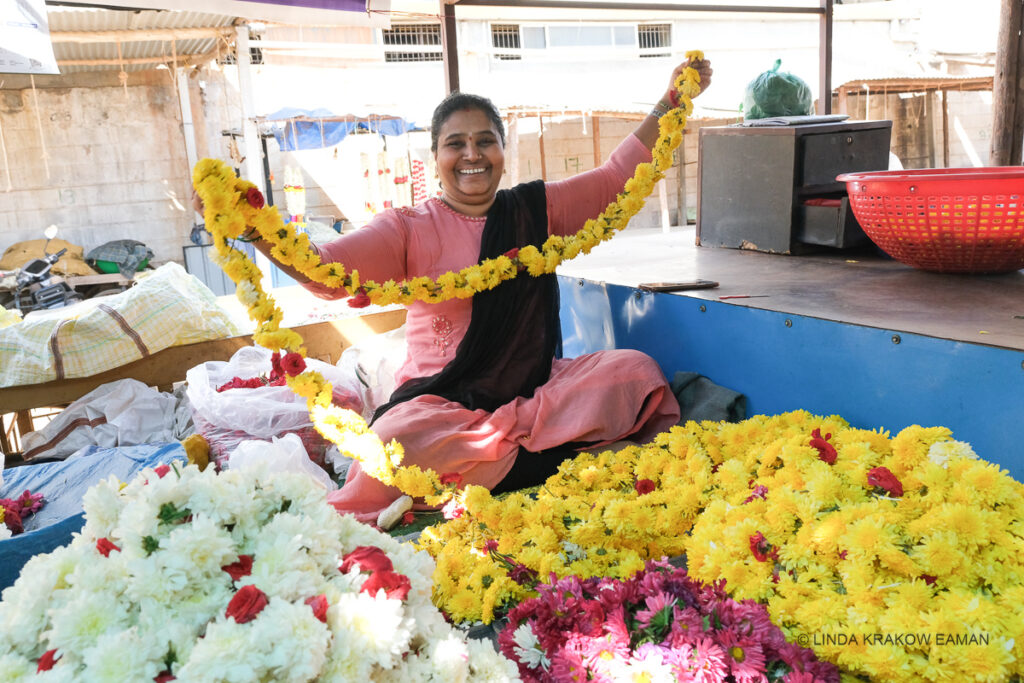 A woman in a pink saree is sitting cross legged in front of piles of yello, pink, and white chrysanthemum blossoms. She is smiling and holding a long string of yellow chrysanthemums, with an occasional red one for accent, in her outstretched arms. 
