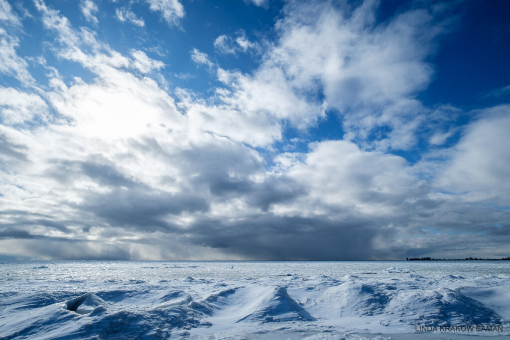 A dramatic scene with mounds of ice and snow in the foreground and icy lake Ontario stretching to the horizon. Big puffy white clouds reach up to a very blue sky. There are darker bands of precipitation where the sky meets the water.